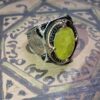 A silver ring with a large, oval, yellow-green gemstone. The ring has intricate filigree and gold-colored accents around the stone and on the band. The ring is set against a patterned background. — Khodamdjin