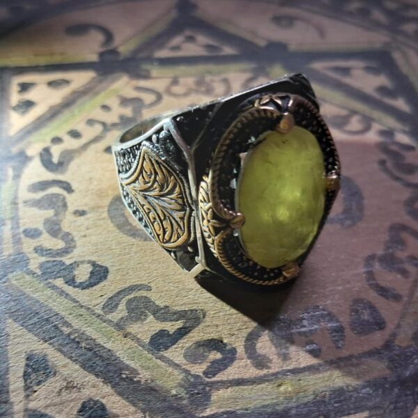 A silver ring with a large, oval, yellow-green gemstone. The ring has intricate gold-colored filigree and dark accents. The setting is ornate, with detailed patterns on the sides. — Khodamdjin