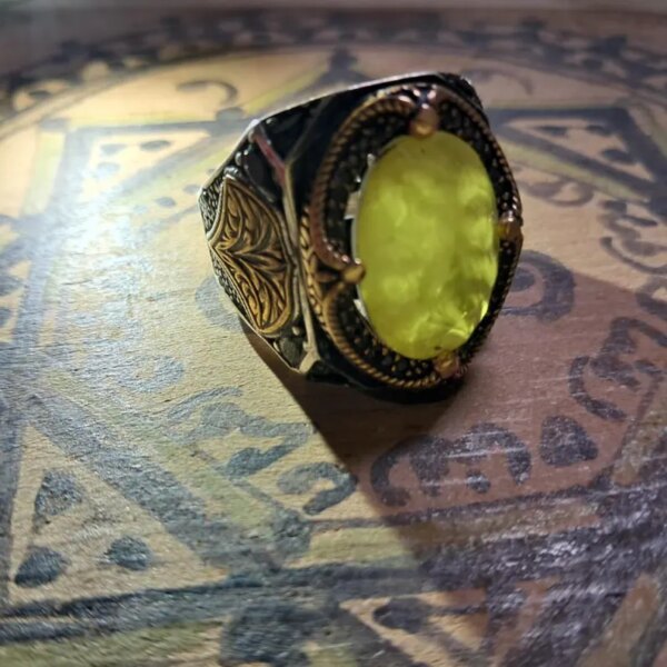 A silver ring with gold accents and a large, oval, yellow gemstone. The ring has intricate filigree designs on the sides and around the gemstone. The setting is dark, contrasting with the bright stone. — Khodamdjin