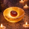 A glowing, translucent orange stone bowl with a smooth, rounded surface. Inside the bowl sits a single, dark reddish-brown, oval-shaped stone. The bowl emits a warm, soft light, highlighting its crystalline texture and natural mineral veins. — Khodamdjin