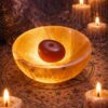 A glowing, translucent orange stone bowl with a smooth, rounded surface. Inside the bowl sits a single, dark reddish-brown, oval-shaped stone. The bowl emits a warm, soft light, highlighting its crystalline texture and natural mineral veins. — Khodamdjin