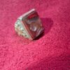 A vintage ring with a rectangular, reddish-brown stone. The stone has a carved design and is set in a silver-colored metal band with ornate detailing. The ring is displayed on a red surface. — Khodamdjin