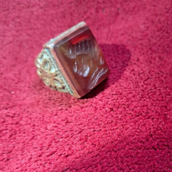 A vintage ring with a rectangular, reddish-brown stone. The stone has a carved design and is set in a silver-colored metal band with ornate detailing. The ring is displayed on a red surface. — Khodamdjin
