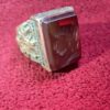 A rectangular gemstone ring with a gold-colored band. The gemstone is a deep reddish-brown color. The band features an ornate, embossed design. The ring is set against a red background. — Khodamdjin
