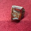 A rectangular, reddish-brown ring with a carved design on the face. The ring has a silver-colored band with an ornate pattern on the sides. The carved design appears to be a profile of a person or figure. — Khodamdjin