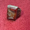 A rectangular, reddish-brown gemstone ring with a detailed carving on the face. The ring has a silver-colored band with an ornate design. The gemstone is set in a silver bezel. — Khodamdjin