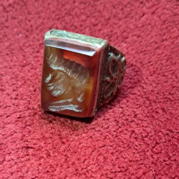 A rectangular, reddish-brown gemstone ring with a detailed carving on the face. The ring has a silver-colored band with an ornate design. The gemstone is set in a silver bezel. — Khodamdjin