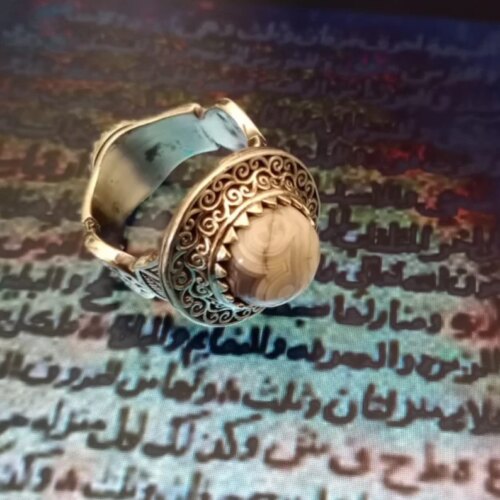A silver-toned metal ring featuring an intricate filigree pattern surrounding a central, smooth, round, beige and brown banded stone cabochon. The band of the ring displays detailed metalwork with a polished finish and a wide design. — Khodamdjin