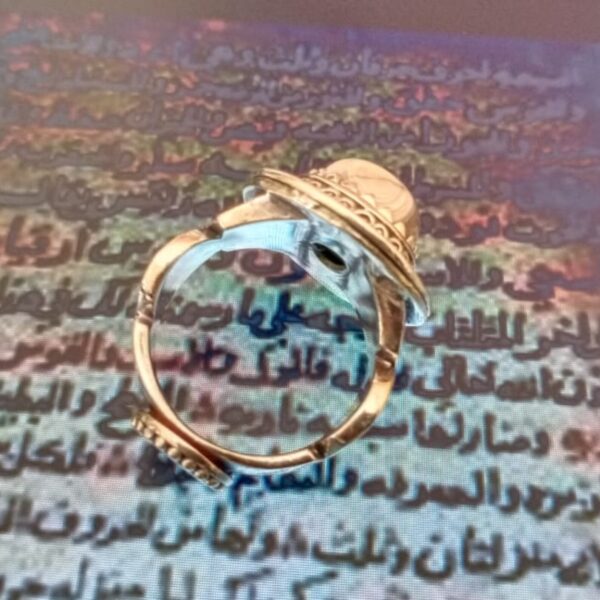 A gold-toned ring featuring a circular band with decorative filigree detailing along the outer edge. The ring has a central setting that holds a smooth, rounded stone, showcasing a metallic finish and intricate metalwork on the shank. — Khodamdjin