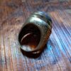 A silver-toned metal ring featuring an oval-shaped, dark, mottled stone set in the center. The band of the ring is wide and decorated with intricate, embossed scrollwork patterns that wrap around the sides of the metal setting. — Khodamdjin