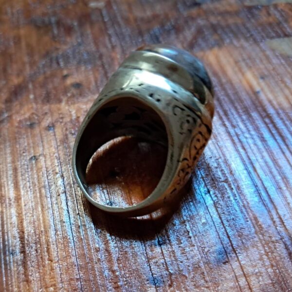 A silver-toned metal ring featuring an oval-shaped, dark, mottled stone set in the center. The band of the ring is wide and decorated with intricate, embossed scrollwork patterns that wrap around the sides of the metal setting. — Khodamdjin