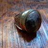 A metal ring featuring a large, rounded, dark brown and grey mottled stone set in a polished band. The sides of the ring are decorated with intricate, engraved scrollwork patterns that wrap around the metal shank. — Khodamdjin