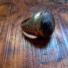 A metal ring featuring a large, smooth, dark gray and black mottled stone set in a polished band. The sides of the ring are decorated with intricate, engraved scrollwork patterns that wrap around the metal shank. — Khodamdjin