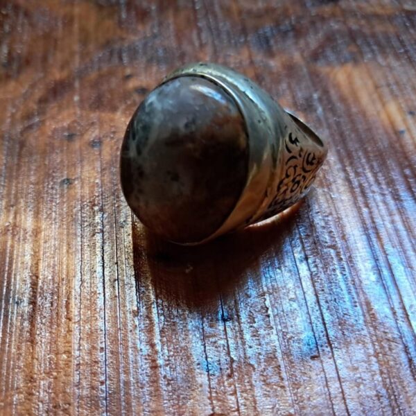 A silver-toned metal ring featuring a large, oval-shaped, polished brown and grey mottled stone cabochon. The band of the ring displays intricate, dark, engraved scrollwork patterns along its outer surface. — Khodamdjin
