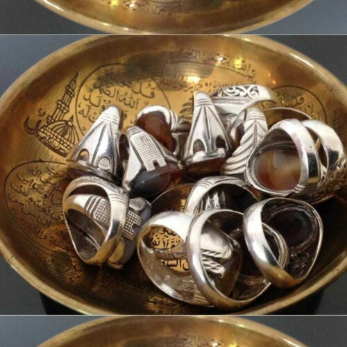 A collection of silver-toned rings with smooth, oval-shaped brown stones resting inside a round, metallic gold-colored bowl. The bowl features intricate engraved Arabic calligraphy and a detailed illustration of a mosque dome. — Khodamdjin