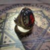 A silver-toned metal ring featuring an oval-shaped, faceted deep red gemstone at its center. The band and setting are decorated with intricate, embossed patterns and a border of small, orange-colored stones surrounding the center gem. — Khodamdjin