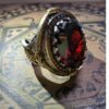 A silver-toned metal ring featuring an oval-shaped, deep red faceted gemstone centerpiece. The band is decorated with intricate, embossed filigree patterns and is surrounded by a border of small, dark orange stones. — Khodamdjin