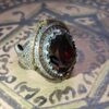 A silver-toned ring featuring an oval-cut dark red gemstone at the center. The setting is decorated with intricate metalwork, a border of small orange stones, and gold-toned accents along the sides of the band and the bezel. — Khodamdjin