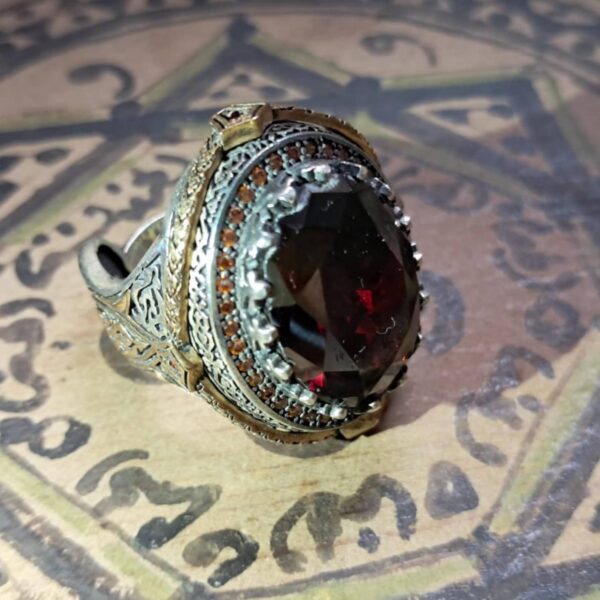 A silver-toned ring featuring an oval-cut dark red gemstone at the center. The setting is decorated with intricate metalwork, a border of small orange stones, and gold-toned accents along the sides of the band and the bezel. — Khodamdjin