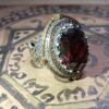 A silver and gold-toned ring featuring a large, oval-cut dark red gemstone at the center. The setting is surrounded by a border of small orange stones and intricate metal filigree work along the band, showcasing detailed engravings. — Khodamdjin