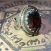 A silver-toned ring featuring a large, oval-cut dark red gemstone at its center. The setting is surrounded by a border of small, amber-colored stones and intricate, textured metalwork along the band and sides of the ring head. — Khodamdjin