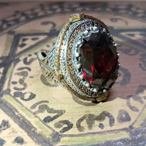 A silver-toned ring featuring a large, oval-cut dark red gemstone at its center. The setting is surrounded by a border of small, amber-colored stones and intricate, textured metalwork along the band and sides of the ring head. — Khodamdjin