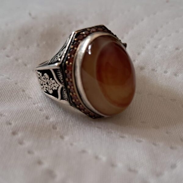 A silver ring featuring a large, oval, smooth, light brown agate stone set in a bezel. The metal band is decorated with intricate black-filled filigree patterns and a border of small, dark, reddish-brown stones around the central setting. — Khodamdjin