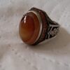 A silver ring featuring a large, smooth, oval-shaped amber-colored stone set in the center. The band is decorated with intricate black oxidized metal engravings and a border of small, reddish-orange stones surrounding the main gem. — Khodamdjin
