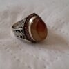 A silver ring featuring a large, oval-shaped, smooth, brown and tan banded agate stone set in a bezel. The metal band is decorated with intricate black-filled engravings and a border of small, reddish-brown stones around the center. — Khodamdjin