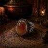 A silver ring featuring a large, smooth, oval-shaped orange and cream-colored agate stone. The silver band is decorated with intricate black engraved patterns and a row of small, dark red stones set along the upper edge of the setting. — Khodamdjin