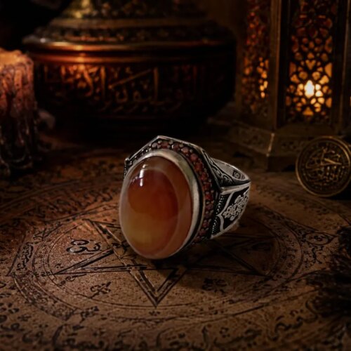 A silver ring featuring a large, smooth, oval-shaped orange and cream-colored agate stone. The silver band is decorated with intricate black engraved patterns and a row of small, dark red stones set along the upper edge of the setting. — Khodamdjin