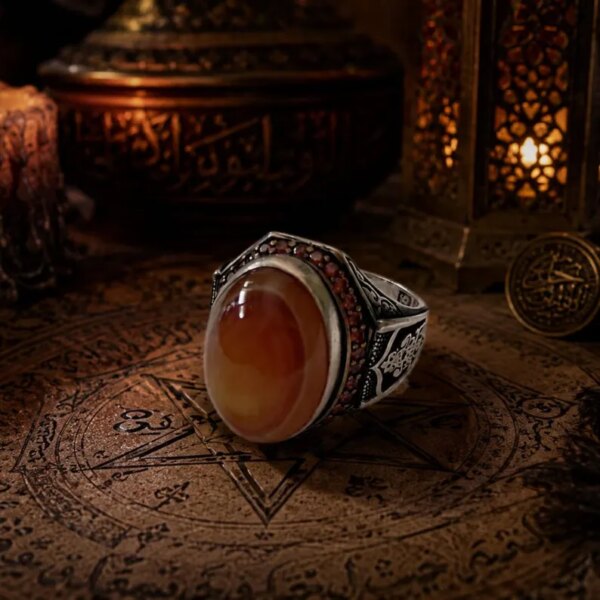 A silver ring featuring a large, smooth, oval-shaped orange and cream-colored agate stone. The silver band is decorated with intricate black engraved patterns and a row of small, dark red stones set along the upper edge of the setting. — Khodamdjin