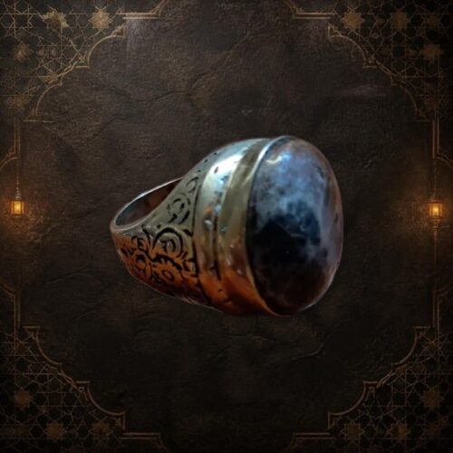 A metal ring featuring a large, smooth, dark gray and black mottled stone set in a polished band. The sides of the ring are decorated with intricate, engraved scrollwork patterns that wrap around the metal shank. — Khodamdjin