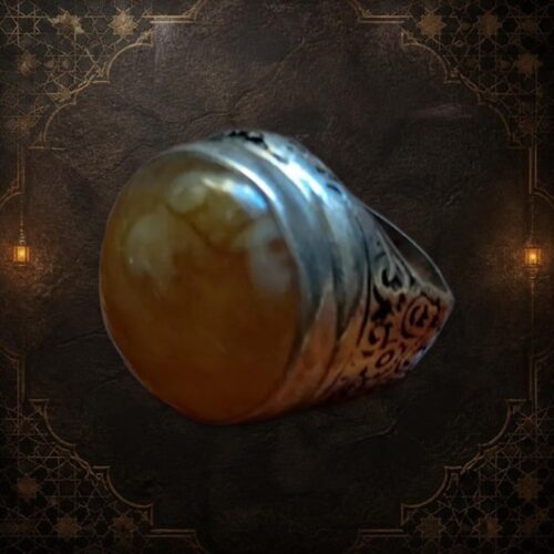 A silver ring featuring a large, smooth, round, amber-colored stone set in a metal band. The band is decorated with intricate, dark, engraved scrollwork patterns that wrap around the sides of the ring's setting. — Khodamdjin