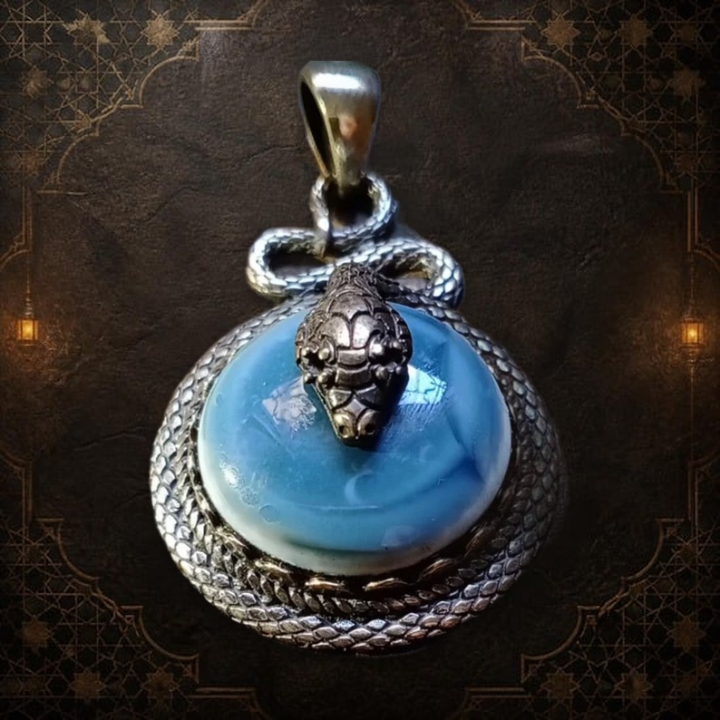 A circular pendant featuring a smooth, light blue cabochon stone set within a textured, silver-toned metal frame that resembles coiled snake scales. A detailed, three-dimensional silver-toned snake head rests atop the stone. — Khodamdjin