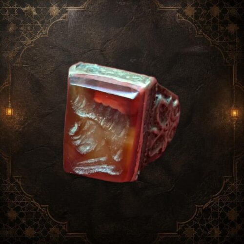 A rectangular, reddish-brown gemstone ring with a detailed carving on the face. The ring has a silver-colored band with an ornate design. The gemstone is set in a silver bezel. — Khodamdjin