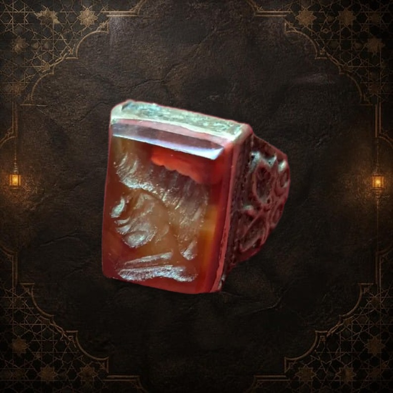 A rectangular, reddish-brown gemstone ring with a detailed carving on the face. The ring has a silver-colored band with an ornate design. The gemstone is set in a silver bezel. — Khodamdjin
