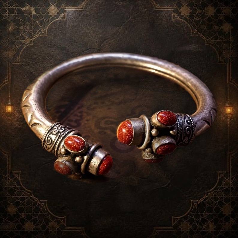 A silver-toned metal cuff bracelet featuring an open design with engraved patterns along the band. The two ends are decorated with multiple oval-shaped, reddish-brown glittery stones set in metal casings, creating a symmetrical appearance. — Khodamdjin